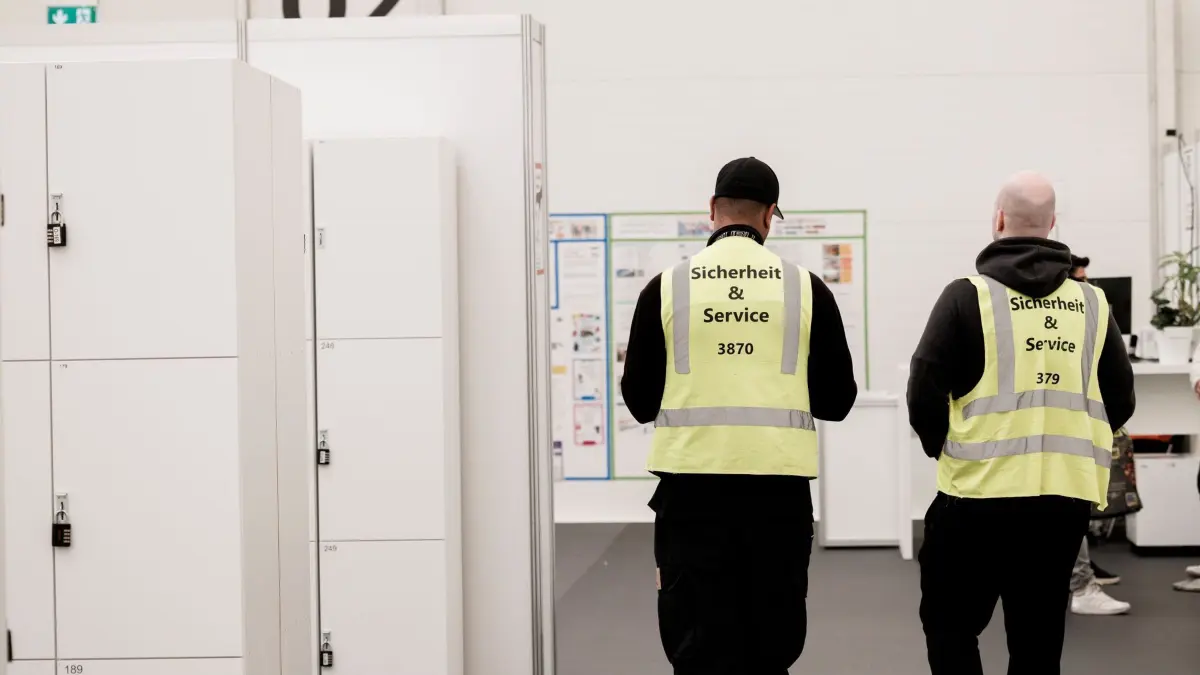 Ankunftszentrum Tegel: 28.05.2024, Berlin: Zwei Mitarbeiter einer Sicherheitsfirma gehen durch den großen Aufenthaltsraum im Unterkunftskomplex im Ankunftszentrum Tegel. Foto: Carsten Koall/dpa +++ dpa-Bildfunk +++