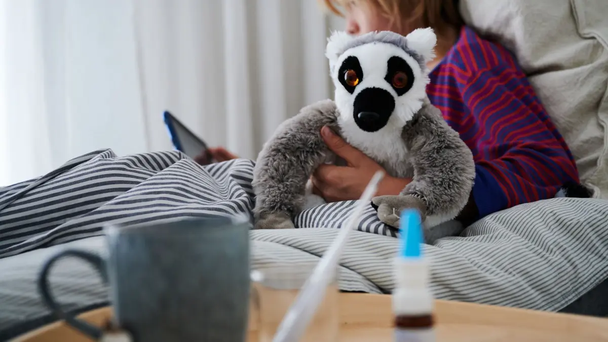 Symbolbild - krankes Kind: ARCHIV - 11.09.2021, Berlin: ILLUSTRATION - Ein Fieberthermometer, Nasenspray und eine Tasse Tee stehen vor einem Bett, in dem ein krankes Kind mit einem Kuscheltier sitzt und auf ein Tablet schaut. (zu dpa: «Mykoplasmen-Infektionen beschäftigen Kinderärzte») Foto: Annette Riedl/dpa +++ dpa-Bildfunk +++