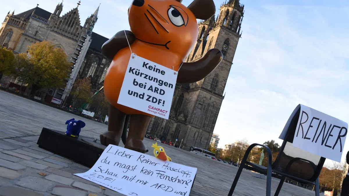 Entführte Figur der „Sendung mit der Maus“ steht in Magdeburg vo: 17.10.2024, Sachsen-Anhalt, Magdeburg: Die von Aktivisten von «Campact e.V.» in Köln entführte Maus aus der «Sendung mit der Maus» steht mit Schildern «Keine Kürzungen bei ARD und ZDF!» und «Lieber Reiner Haseloff, du magst kein Fernsehen? Schau mit mir ARD und ZDF! Kommst Du?» auf dem Domplatz. Die Aktivisten reisen mit der Maus in mehrere deutsche Städte, um ein Zeichen gegen die drastischen Kürzungen im Informations- und Bildungsangebot des öffentlich-rechtlichen Rundfunks (ÖRR) zu setzen. Foto: Simon Kremer/dpa +++ dpa-Bildfunk +++