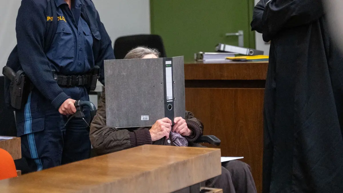Auftakt Mordprozess gegen eine 90-Jährige: 17.04.2025, Bayern, München: Eine 90 Jahre alte Angeklagte wird von einer Polizistin in einem Rollstuhl in einen Sitzungssaal im Landgericht München I geschoben und hält sich einen Aktenordner vor das Gesicht. Im Landgericht München I findet der Auftakt zum Mordprozess gegen eine 90-Jährige, die ihre 77 Jahre alte Freundin mit verschiedenen Küchenutensilien umgebracht haben soll, statt. Die Frau ist wegen Mordes aus Heimtücke und niedriger Beweggründe angeklagt. Foto: Leonie Asendorpf/dpa +++ dpa-Bildfunk +++
