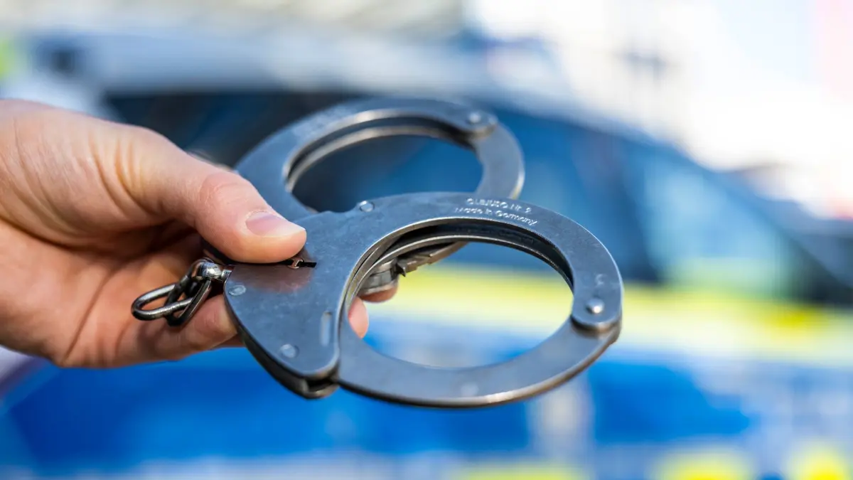 Handschellen vor Polizeiwagen - Symbolbild: ARCHIV - 28.09.2021, Nordrhein-Westfalen, Gütersloh: Eine Hand hält Handschellen vor einen Streifenwagen der Polizei. (zu dpa: «Polizei nimmt mutmaßlichen Serien-Exhibitionisten fest») Foto: David Inderlied/dpa +++ dpa-Bildfunk +++