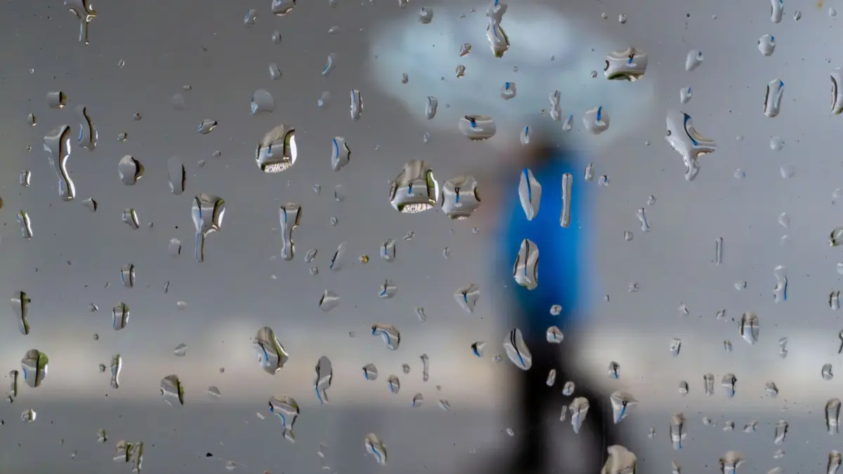 Wetter in Bayern: 30.03.2026, Bayern, Untermerzbach: Ein Junge spiegelt sich in zahlreichen Regentropfen an einer Fensterscheibe, während er mit einem Regenschirm eine Straße entlanggeht. Die Osterferien in Bayern beginnen trist. Noch bis zur Wochenmitte erwartet der Deutsche Wetterdienst Regen, Wind und Schnee in Bayern. Foto: Pia Bayer/dpa +++ dpa-Bildfunk +++