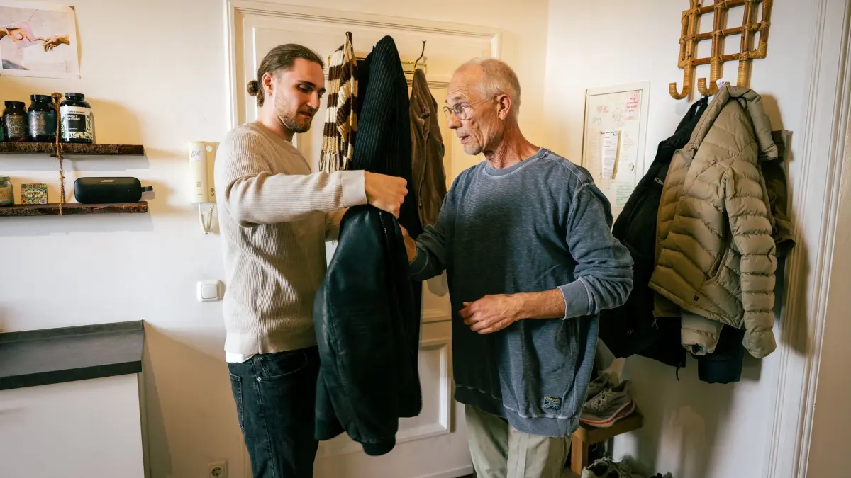 Ein junger Mann hilft einem Senior die Jacke anzuziehen: ILLUSTRATION - Hilfe annehmen: Manchmal braucht es dafür Überwindung und offene Gespräche. (zu dpa: «Intime Momente: Scham in der Pflege ernst nehmen und lindern») Foto: Nico Tapia/dpa-tmn - Honorarfrei nur für Bezieher des dpa-Themendienstes +++ dpa-Themendienst +++