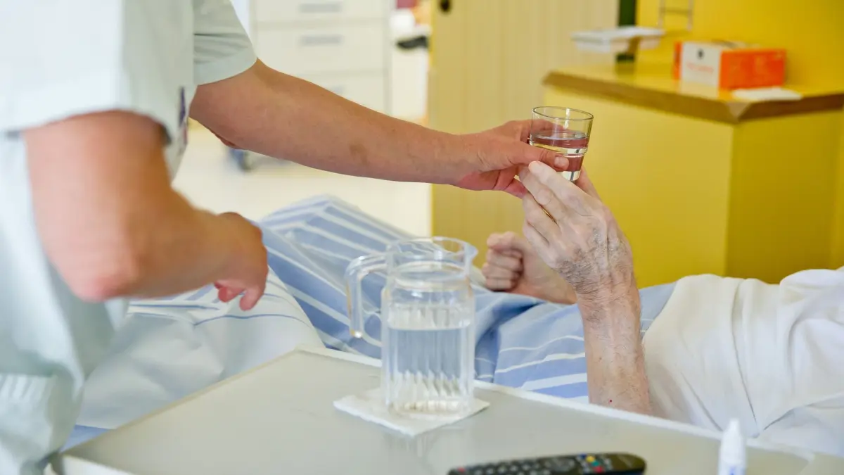 Pflegebedürftiger Mensch: ARCHIV - 01.07.2015, Bayern, Nürnberg: Eine Krankenpflegerin reicht einem Patienten ein Glas Wasser. (zu dpa: «Wie gut sind Pflegeheime und Kliniken gegen Hitze gerüstet?») Foto: Daniel Karmann/dpa +++ dpa-Bildfunk +++