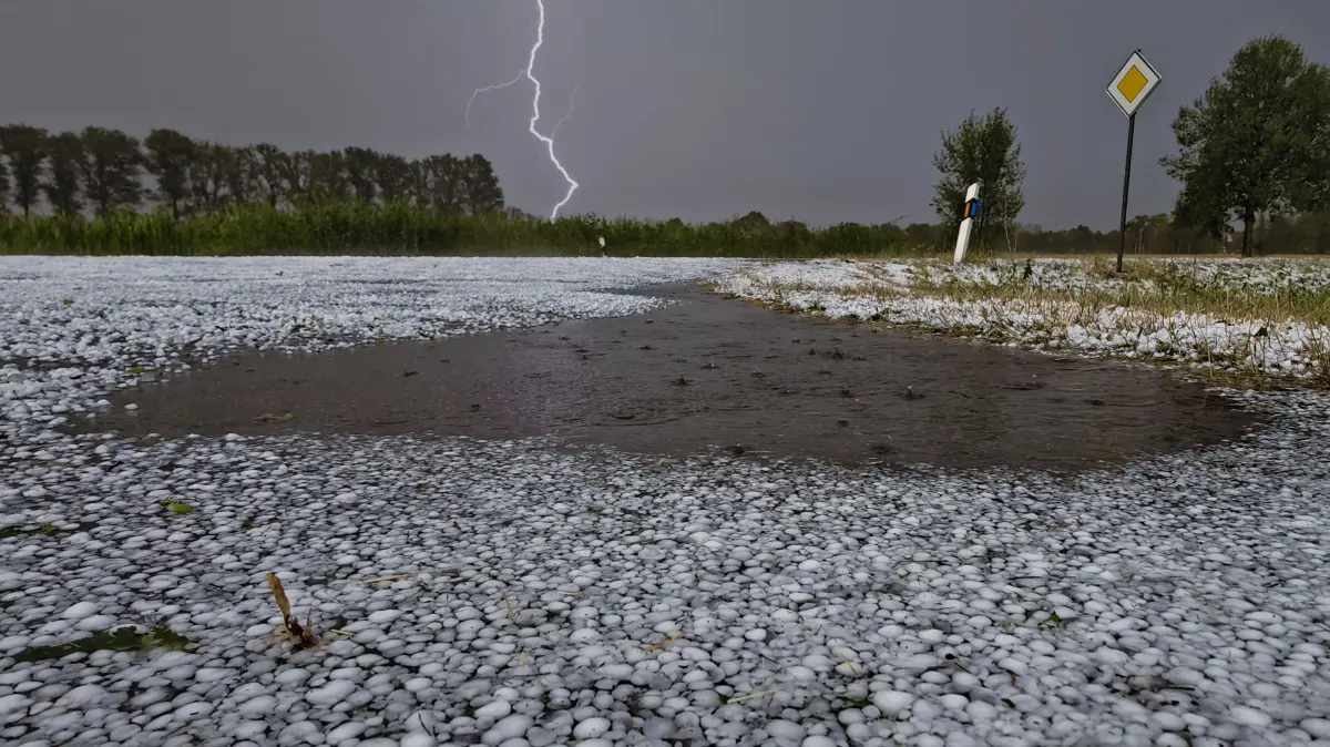 Hagelkörner liegen auf einer Fahrbahn bei Wagenfeld in Niedersachsen, während sich ein Blitz im Hintergrund entlädt. Schwere Unwetter sind über Teilen Niedersachsens niedergegangen.