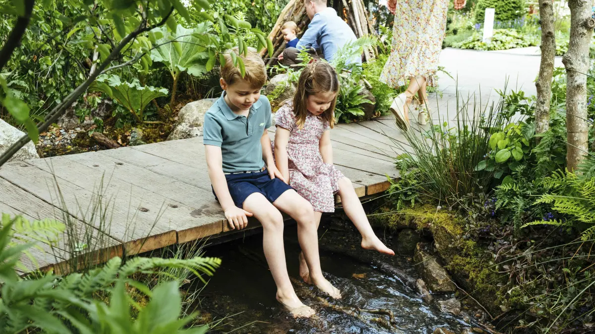Der britische Prinz George und seine Schwester, Prinzessin Charlotte, sitzen auf einem Steg über einen Bach auf dem Gelände des von Adam White und Andree Davies mitgestalteten Garten vor der RHS Chelsea Flower Show, während im Hintergrund Prinz William (r) mit Prinz Louis zu sehen ist.