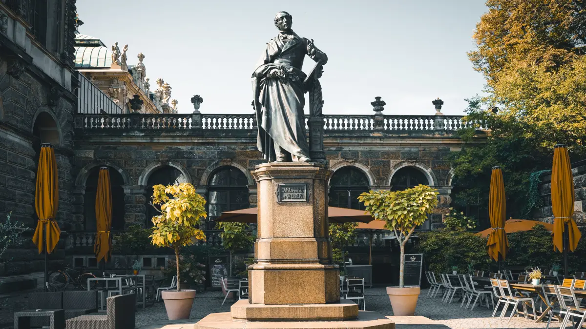 Das Carl-Maria-von-Weber-Denkmal befindet sich am Dresdner Theaterplatz mit Blick auf die Semperoper. Geschaffen vom Dresdner Bildhauer Ernst Rietschel, wurde das Denkmal im Jahr 1861 nach langer Planungszeit feierlich enthüllt. Finanziert durch Spenden und Beiträge des sächsischen Hofes, stand es zunächst vor dem damaligen Hotel Bellevue. In den 1950er-Jahren erhielt das Denkmal seinen heutigen Standort am Theaterplatz. Bis heute erinnert es an den Komponisten Carl Maria von Weber, dessen Musik eng mit Dresden verbunden ist und an der Semperoper regelmäßig aufgeführt wird. Foto: Michael R. Hennig (DML-BY)