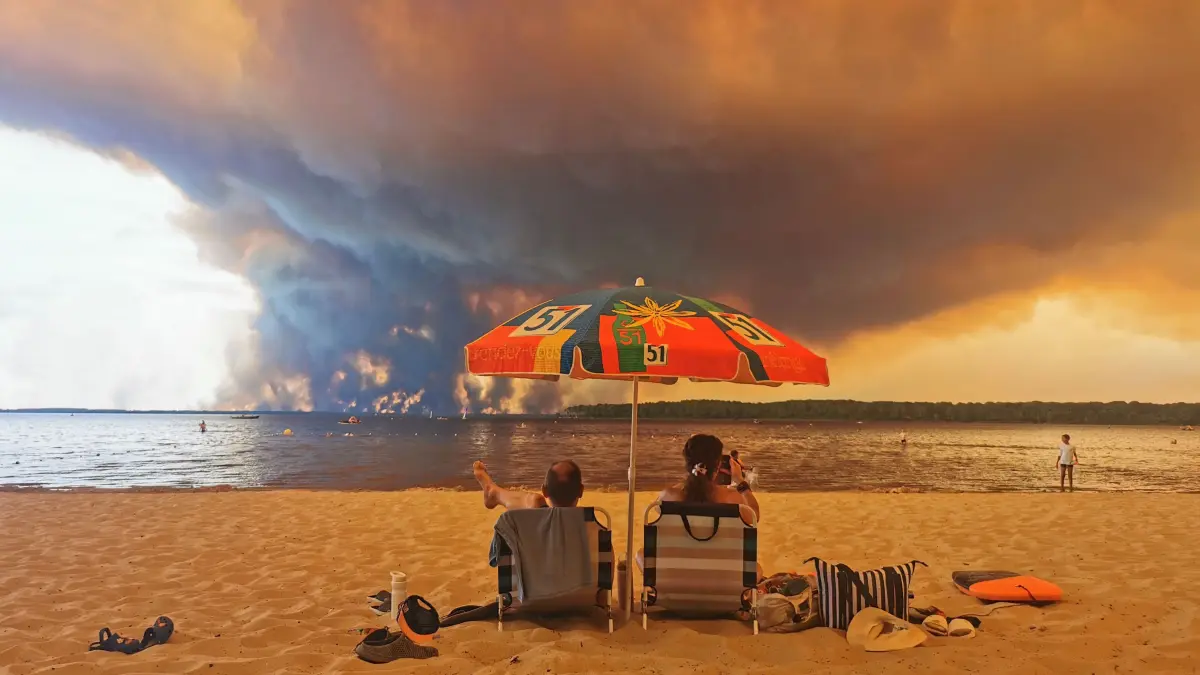 FRANCE-ENVIRONMENT-CLIMATE-WEATHER-WILDFIRE: Beach goers sit on the beach of Moutchic in Lacanau, southwestern France on July 24, 2026, as clouds of smoke rise into the sky from a wildfire. The fire raging in the northern part of the Arcachon Basin in Gironde, southwest France, has swept through more than 3,400 hectares, according to the latest figures released by the prefecture. More than 20,000 residents and tourists have been evacuated from this part already devastated by flames in 2022. Scientists have long warned that climate change caused by human use of fossil fuels will make periods of drought more intense and longer-lasting, creating the ideal conditions for wildfires. (Photo by Maximilien LAMY / AFP)