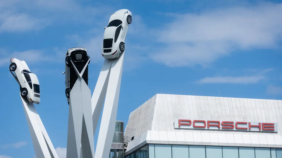 Porsche Werk Zuffenhausen: 27.07.2025, Baden-Württemberg, Stuttgart: Während das Porsche Logo auf einem Gebäude am Firmensitz in Stuttgart-Zuffenhausen zu sehen ist, ragt die „Inspiration 911“ Installation, bestehend aus drei Porsche 911er Modellen in die Höhe. Foto: Christoph Schmidt/dpa +++ dpa-Bildfunk +++