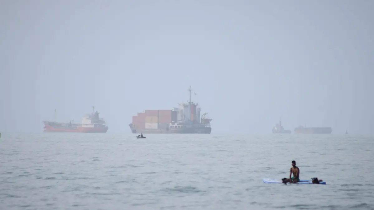 Iran-Krieg - Straße von Hormus: ARCHIV - 27.07.2026, Iran, Bandar Abbas: Ein Mann sitzt auf einem Surfbrett, während ein Containerschiff und andere Handelsschiffe in der Straße von Hormus vor Anker liegen. (zu dpa: «GPS-Störungen: Die unsichtbare Front im Iran-Krieg») Foto: Razieh Poudat/ISNA/AP/dpa +++ dpa-Bildfunk +++
