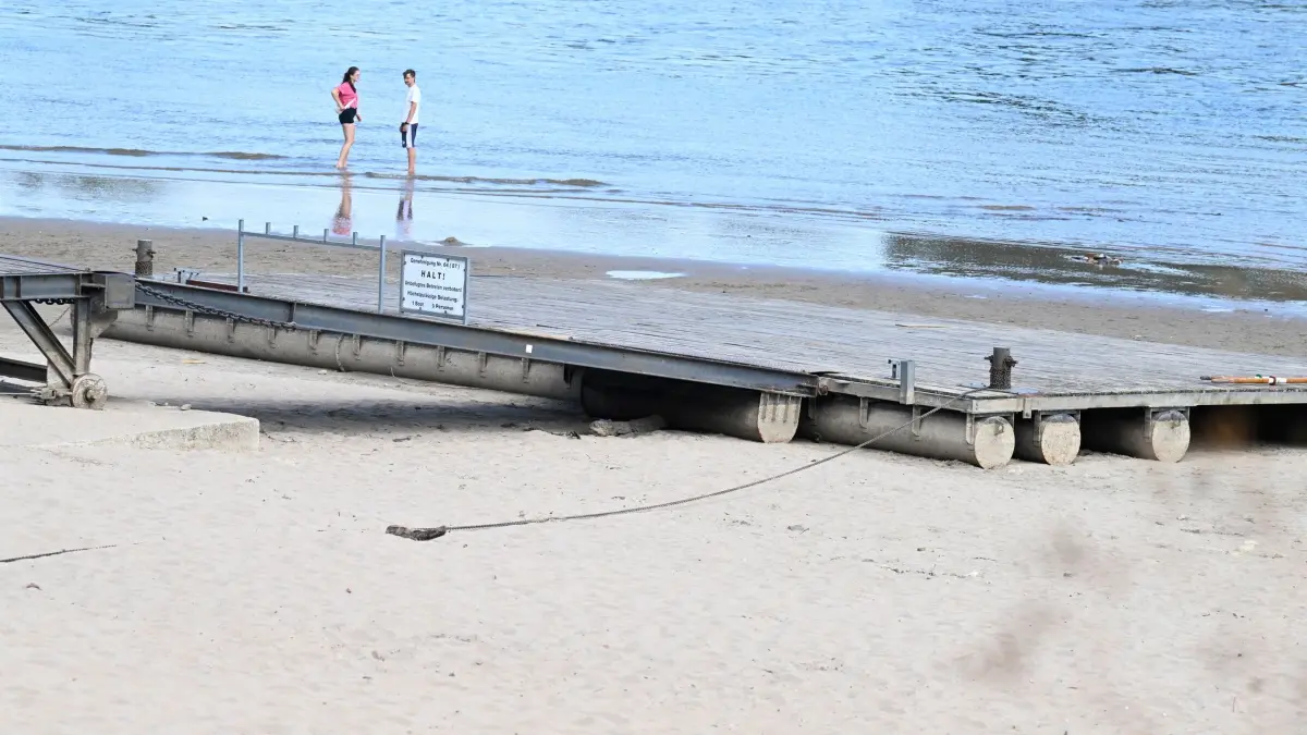 Wetter in Nordrhein-Westfalen: 02.08.2026, Nordrhein-Westfalen, Rodenkirchen: Personen stehen mit den Füßen im flachen Wasser des Rheins, während der Ruderanleger des ortsansässigen Rudervereins aufgrund von Niedrigwasser frei am Ufer liegt. Die Meteorologen rechnen am Ende der nächsten Woche mit Regen, der die Situation beim Niedrigwasser lindern soll. Auch die Temperaturen werden angenehmer erwartet. Foto: Roberto Pfeil/dpa +++ dpa-Bildfunk +++