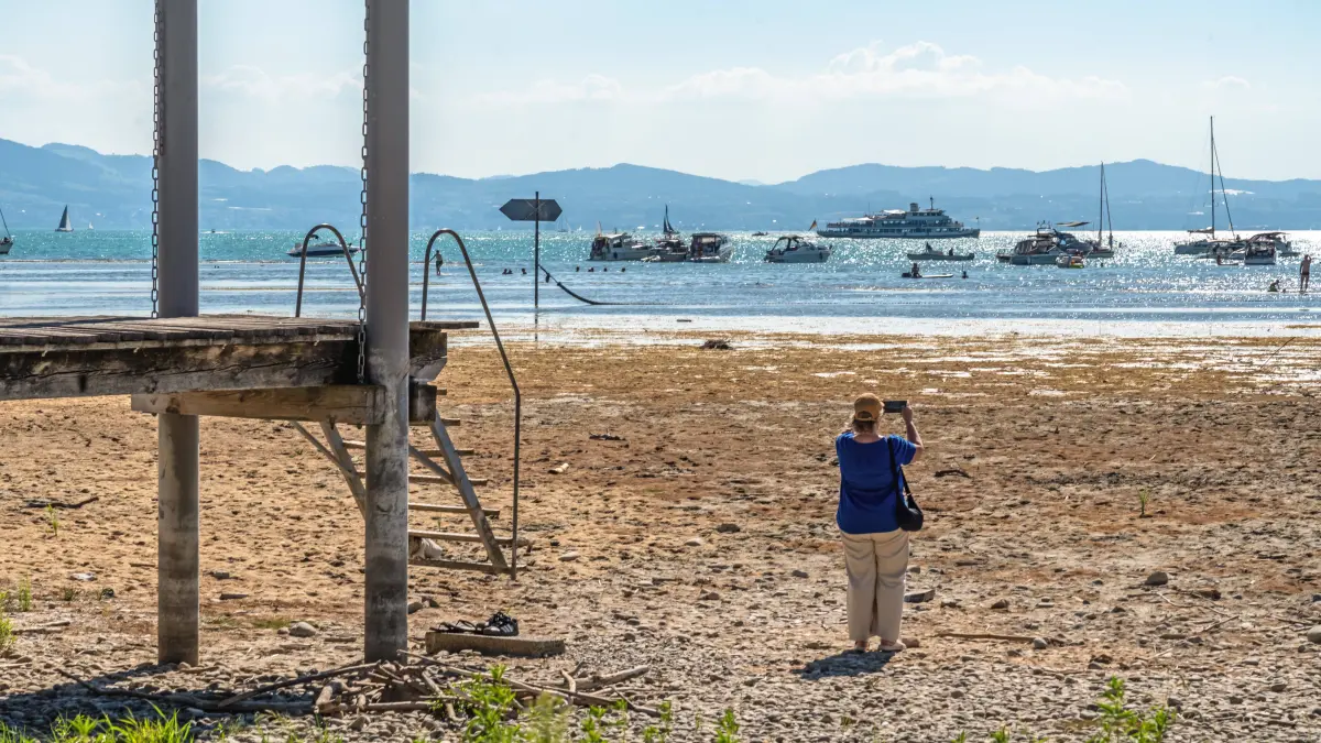 Badesteg ins Trockene, Niedrigwasser am Bodensee, durch Trockenheit freigelegte Uferzone am Bodensee, die Boote liegen weit draußen, die Schifffahrt ist eingeschränkt, Wasserburg, August 2026: Badesteg ins Trockene, Niedrigwasser am Bodensee, durch Trockenheit freigelegte Uferzone am Bodensee, die Boote liegen weit draußen, die Schifffahrt ist eingeschränkt, Wasserburg, August 2026 Deutschland, Wasserburg, 8. August 2026, Badesteg ins Trockene, Niedrigwasser am Bodensee, durch Trockenheit freigelegte Uferzone am Bodensee, ein Badesteg führt ins Trockene, weit entfernt von der Wasserlinie, eine Besucherin fotografiert die weit zurückgewichene Uferlinie, die aktuelle Trockenheit hat den Pegelstand stark sinken lassen, die Boote liegen weit draußen, die Schifffahrt ist eingeschränkt, Niedrigwasser, Wasserstand sehr niedrig, zu wenig Regen, schönes Wetter, Hochsommer, Hitzewelle mit Temperaturen bis 33 Grad, Sommer, Samstagnac