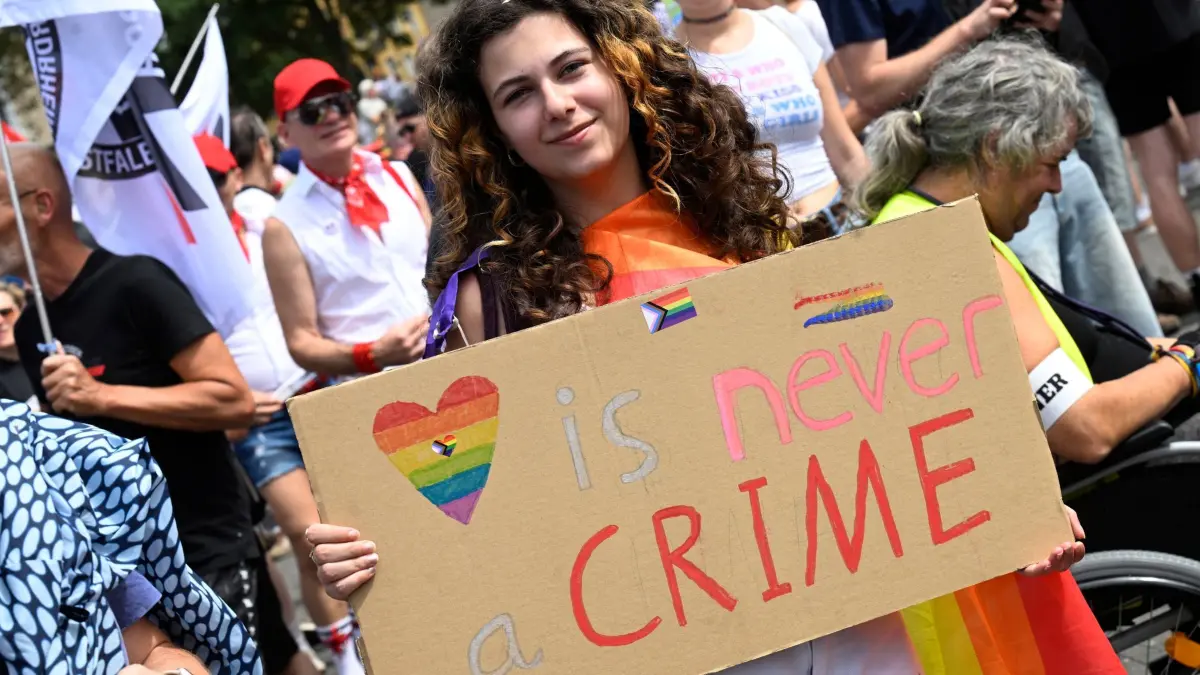 Solidaritätsmarsch für queere Menschen in Köln: 09.08.2026, Nordrhein-Westfalen, Köln: Eine Teilnehmerin des Solidaritätsmarsches unter dem Motto «Köln zeigt Solidarität, Köln zeigt Haltung» von der Deutzer Werft durch die Innenstadt zeigt eine Pappe mit der Aufschrift "is never crime". Mehrere queere Initiativen und Vereine hatten nach dem islamistischen Anschlag am Rande des Berliner CSD dazu aufgerufen. Foto: Roberto Pfeil/dpa +++ dpa-Bildfunk +++