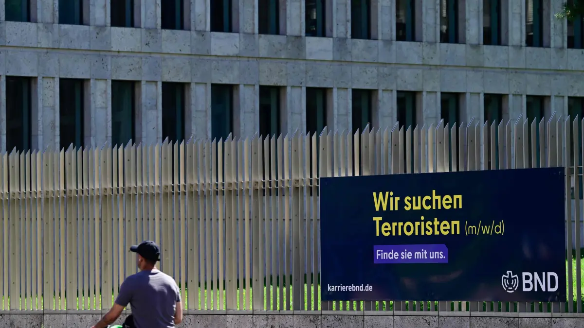 GERMANY-POLITICS-ESPIONAGE-POLICE-GOVERNMENT: A man passes by an job ad reading "We are looking for terrorists" fixed at the fence surrounding the headquarters of the Federal Intelligence Service BND (Bundesnachrichtendienst) in Berlin, on August 12, 2026. The German government signed off on a major overhaul of the country's intelligence agencies to give them broad new powers to conduct covert operations and take action against foreign threats. (Photo by John MACDOUGALL / AFP)
