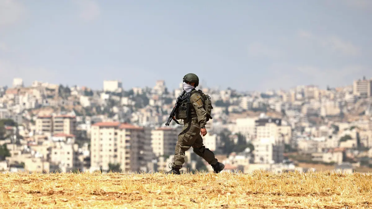 PALESTINIAN-ISRAEL-CONFLICT-WEST BANK-SETTLERS: An Israeli soldier walks through an open field in Beit Sahur, south of Bethlehem, in the occupied West Bank on August 14, 2026. Violence against Palestinians by Israeli settlers in the West Bank has increased dramatically since the October 7, 2023 attack on Israel by Hamas from the Gaza Strip. More than 500,000 Israelis live in settlements in the West Bank, which are illegal under international law, among some three million Palestinians in the territory. (Photo by AHMAD GHARABLI / AFP)