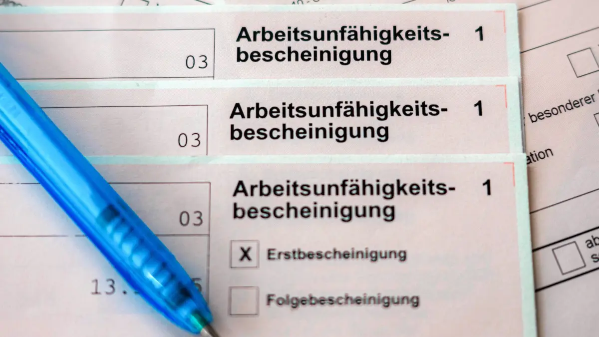 Krankschreibung: ILLUSTRATION - 18.02.2026, Bremen: Mehrere Exemplare einer Arbeitsunfähigkeitsbescheinigung liegen übereinander, daneben ein Kugelschreiber. Berufstätige Versicherte der AOK in Rheinland-Pfalz haben im ersten Halbjahr 2026 krankheitsbedingt etwas weniger am Arbeitsplatz gefehlt. (zu dpa: «AOK-Versicherte in Rheinland-Pfalz melden sich weniger krank») Foto: Sina Schuldt/dpa +++ dpa-Bildfunk +++