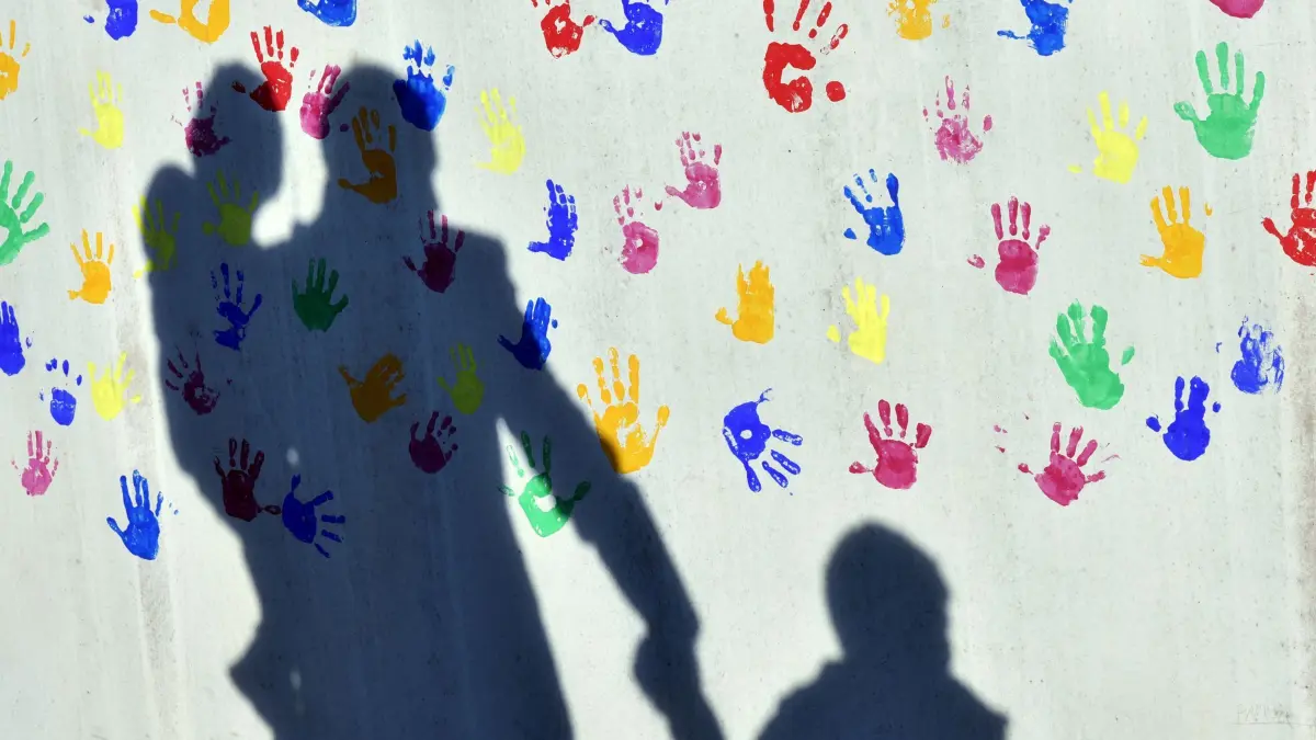 Elterngeld: ARCHIV - 08.04.2015, Bayern, München: Ein Mann mit einem Kind auf dem Arm und einem an der Hand wirft einen Schatten auf eine mit bunten Handabdrücken bemalte Wand einer Kindertagesstätte. (zu dpa: «Elterngeldreform: Mehr Geld und neue Aufteilung geplant») Foto: Peter Kneffel/dpa +++ dpa-Bildfunk +++