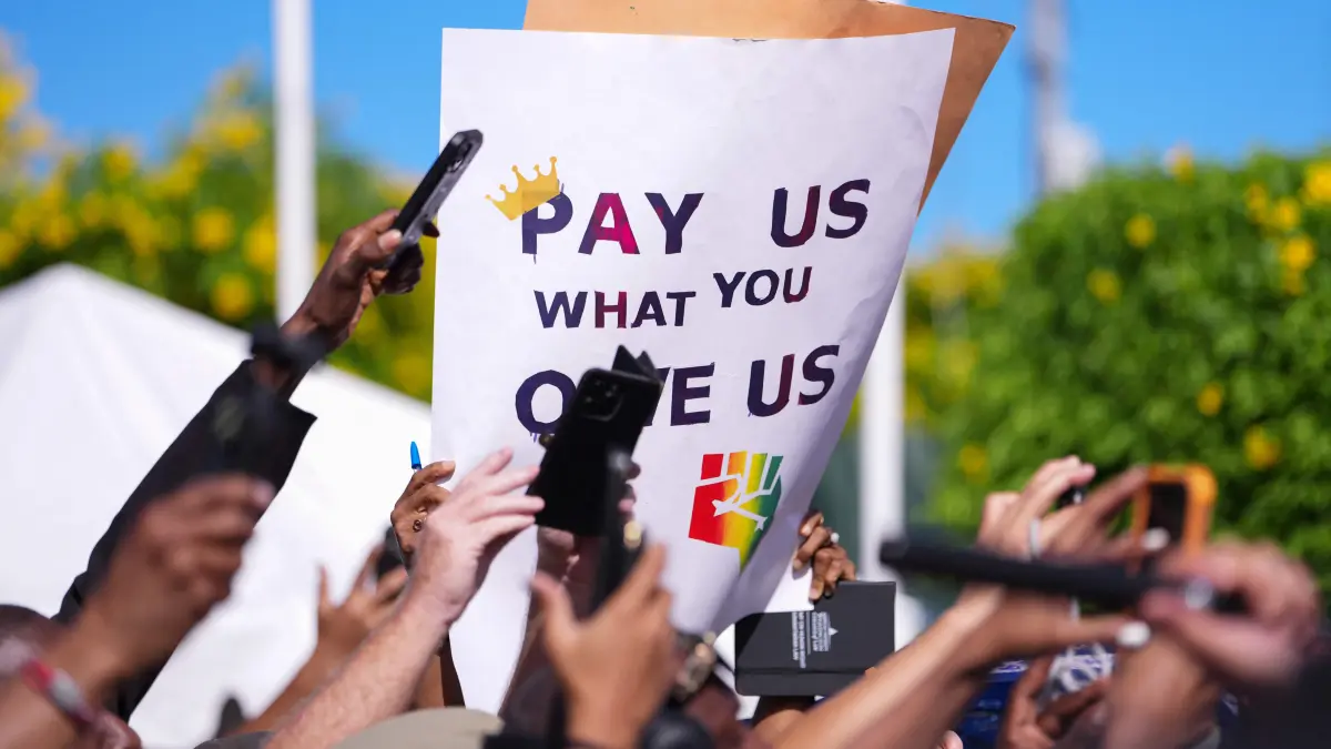 BAHAMAS-BRITAIN-ROYALS: A person in the crowd holds up a sign reading "Pay us what you owe us" as Britain's King Charles III (out of frame) watches a Junkanoo parade in Parliament Square in Nassau, The Bahamas on October 28, 2026. King Charles III's Autumn Tour includes visits to The Bahamas, Guyana and Antigua and Barbuda. (Photo by Ben Stansall / POOL / AFP)